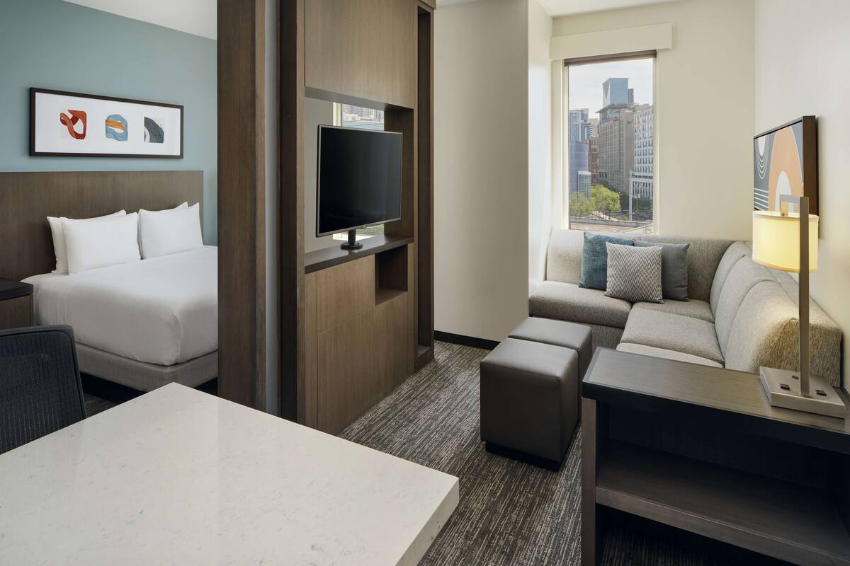 A hotel room with a bed, a TV mounted on a wooden panel, a sofa, a small table, and a window with a city view.