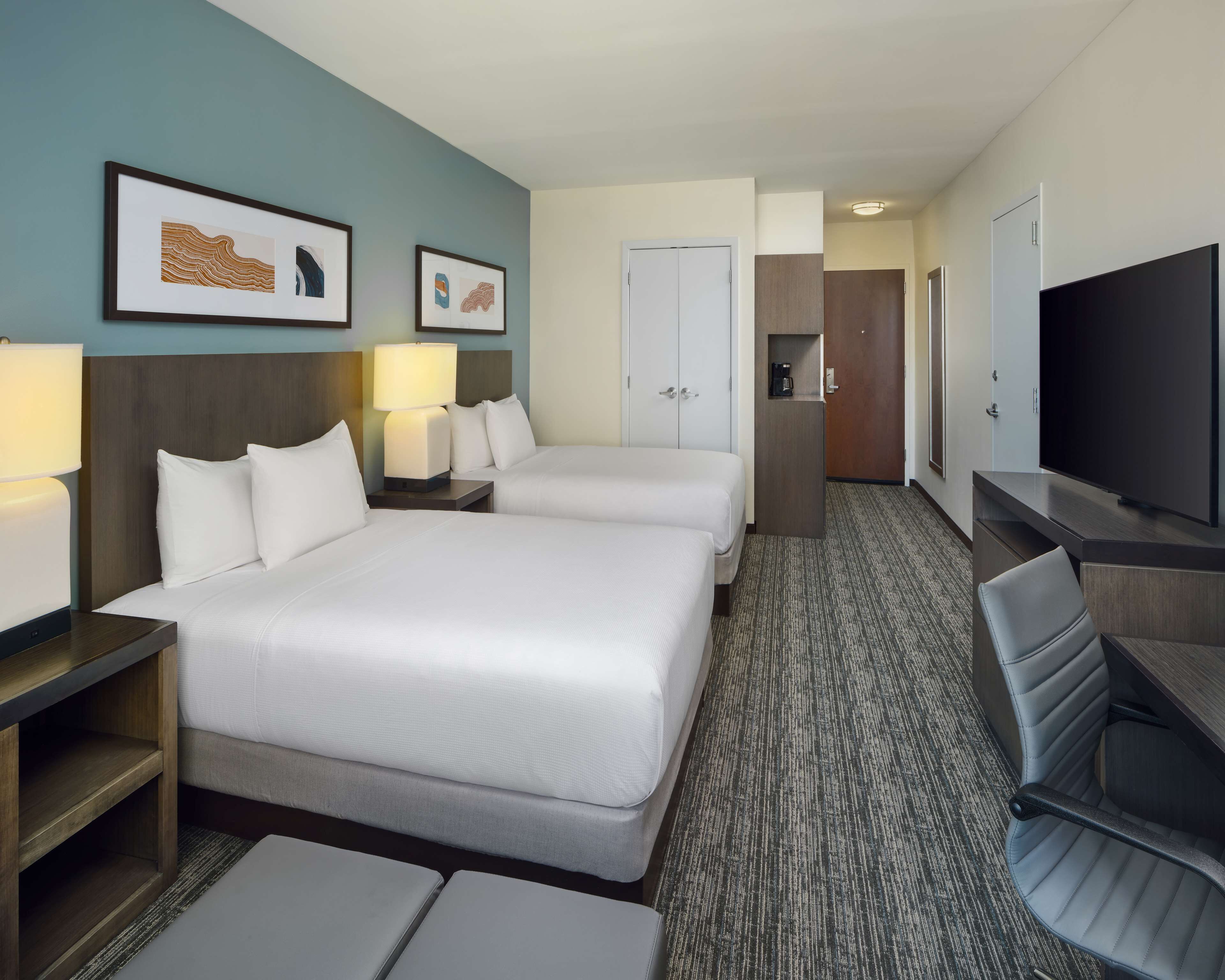 A hotel room with two beds, a desk, a chair, a TV, and a coffee machine.