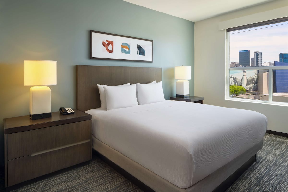 A hotel room with a large bed, two bedside tables with lamps, a framed artwork above the bed, and a window with a city view.