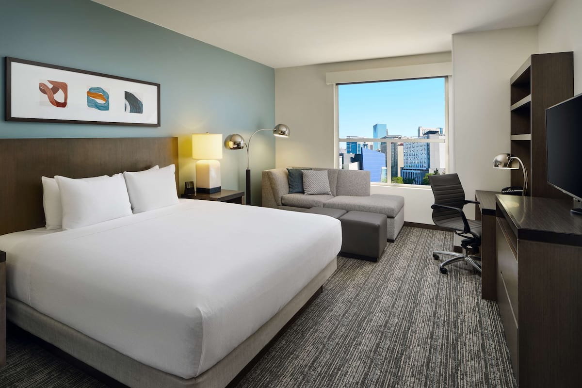 A hotel room with a large bed, a sofa, a desk, and a TV. There is a window with a city view.