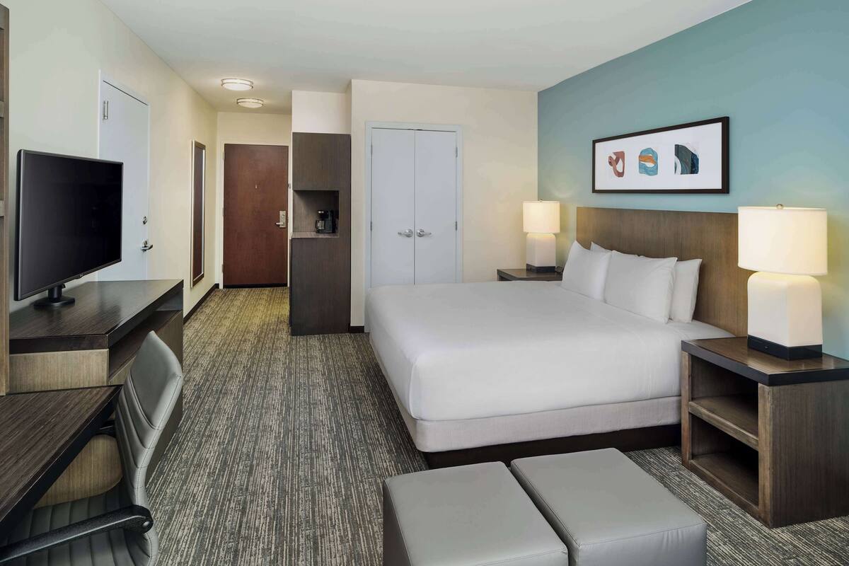A hotel room with a bed, bedside tables, a desk with a TV, a chair, a wardrobe, and a framed picture on the wall.