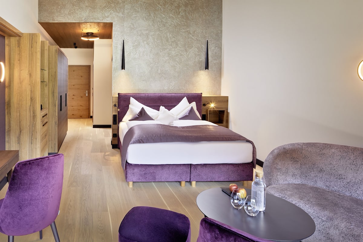 A modern hotel room with a large bed, a sofa, a small table, and a wooden wardrobe.
