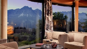 A modern room with a large window offering a mountain view, a sofa, a small table with a book and glasses, and a decorative vase. - Hotel Kaiserhof (Ellmau)