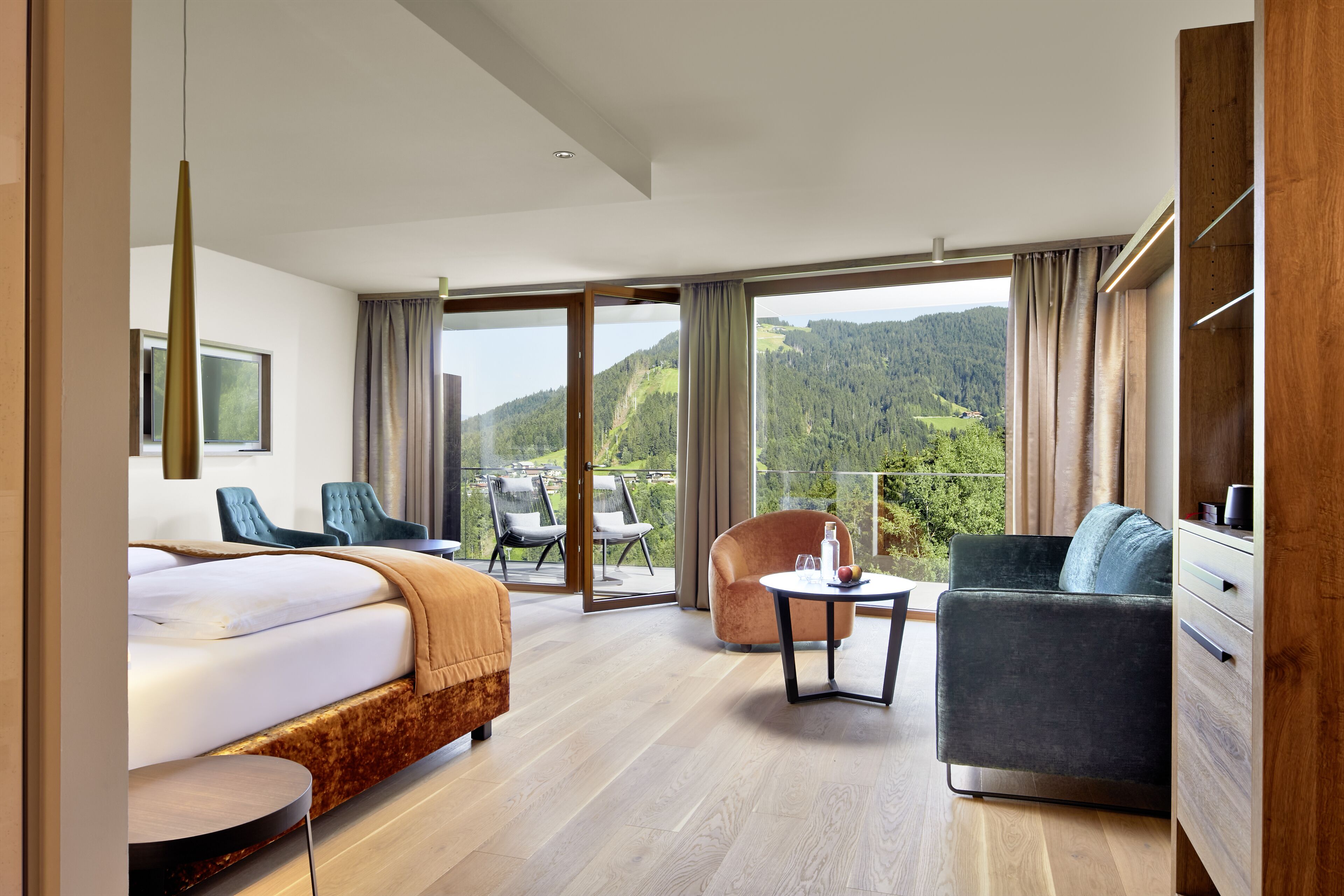 A modern hotel room with a large bed, a sofa, a chair, and a balcony with a scenic mountain view.
