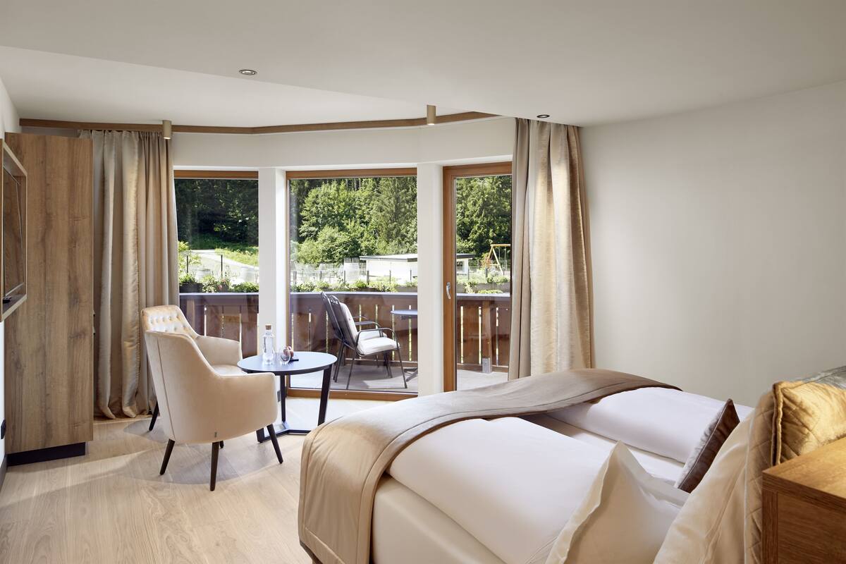 A hotel room with a large bed, a chair, a small table, and a balcony with a view of the outdoors.