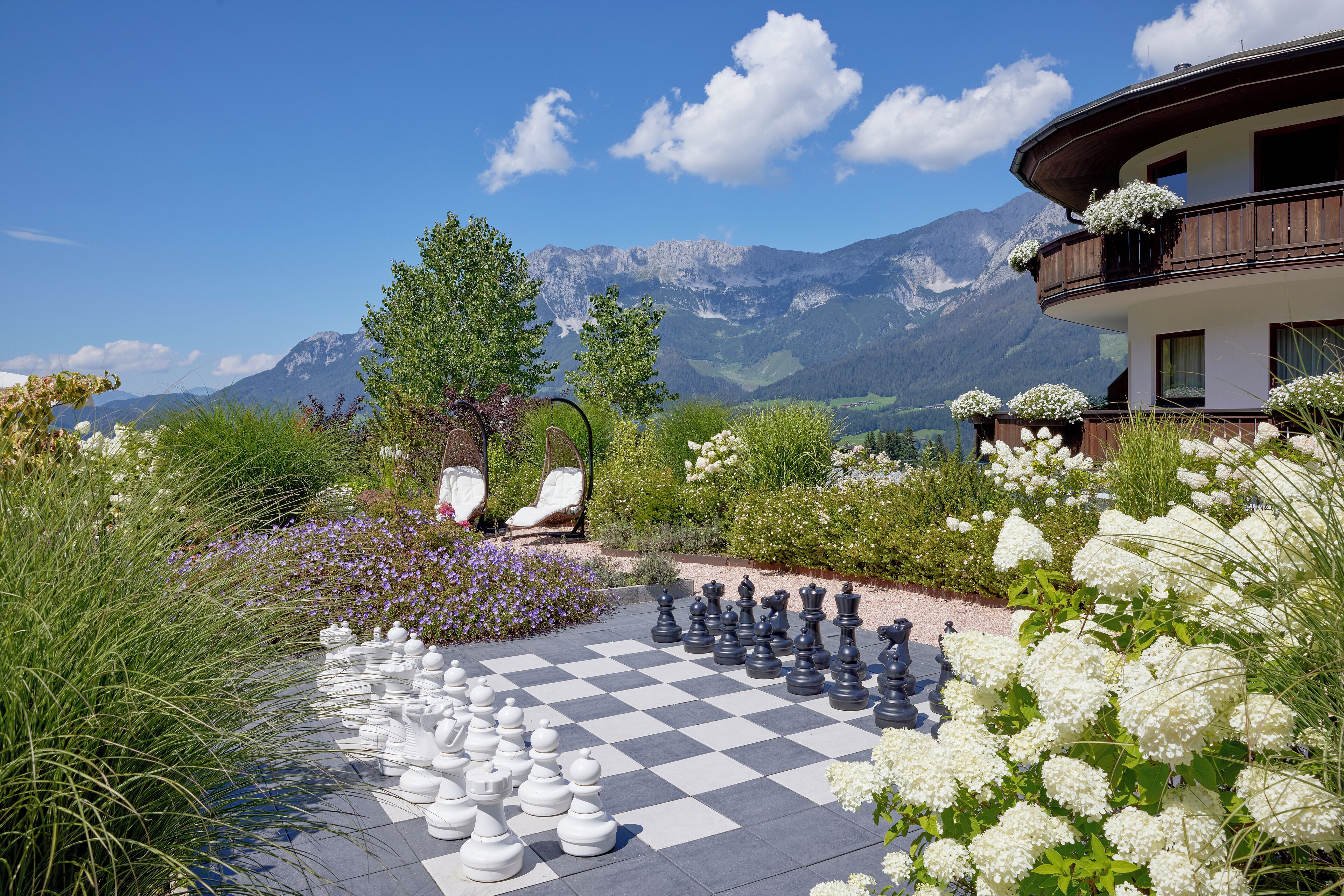 A large outdoor chessboard set up in a garden with mountains in the background.