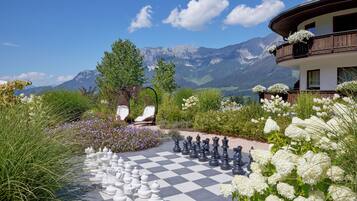 A large outdoor chessboard set up in a garden with mountains in the background.