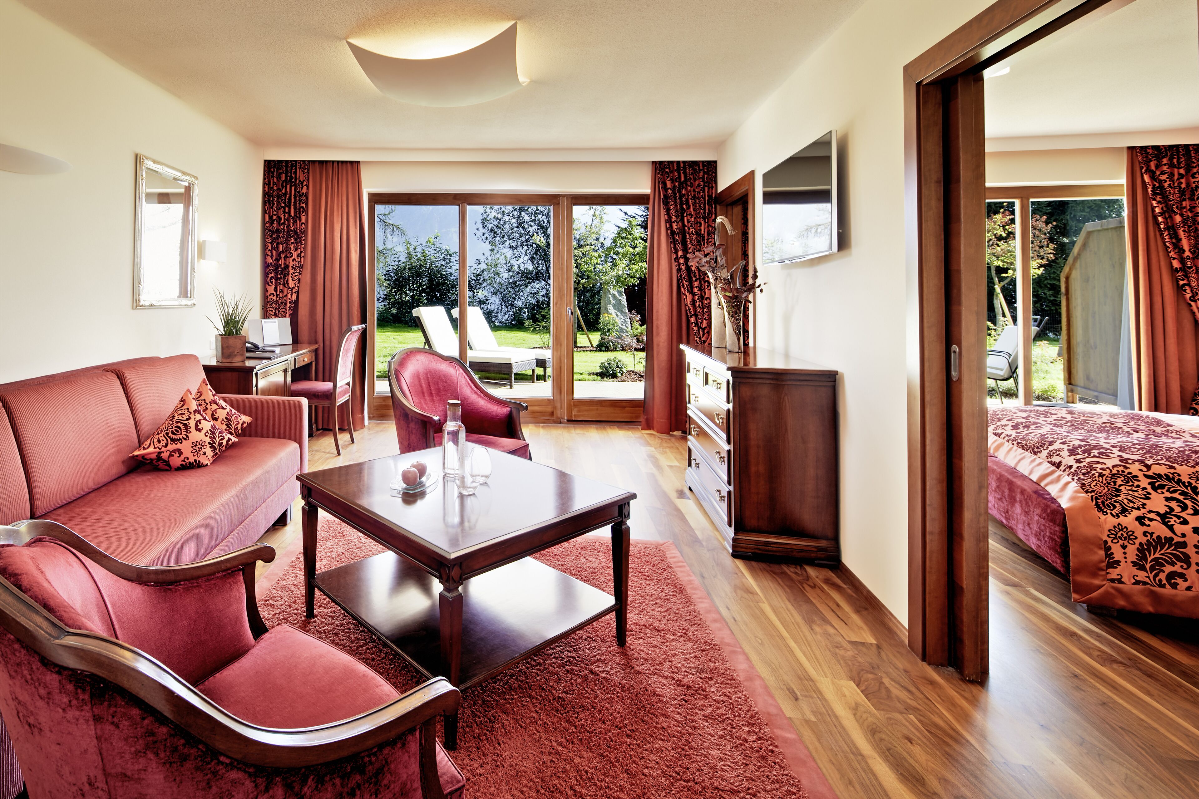 A spacious room with a red sofa, a wooden coffee table, a desk with a chair, and a bed with a patterned bedspread.
