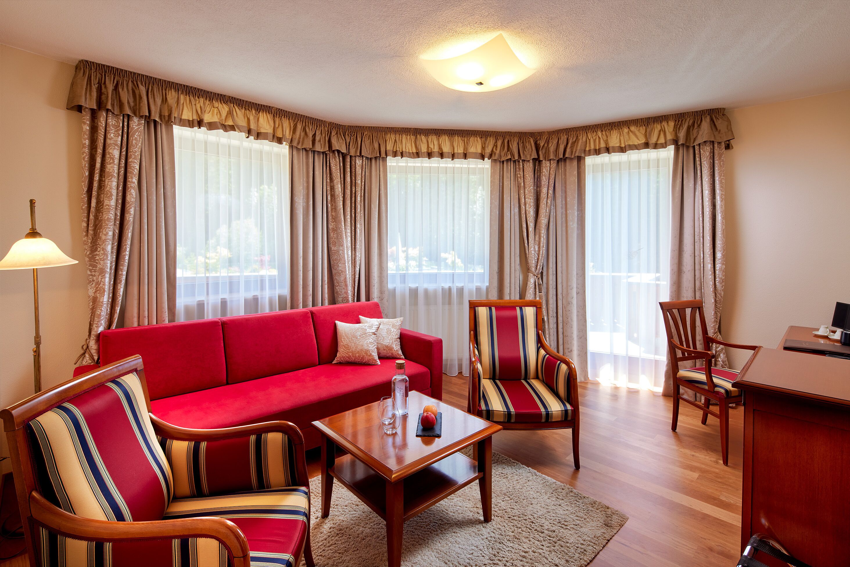 A living room with a red sofa, striped armchairs, a wooden coffee table, and a desk with a lamp.
