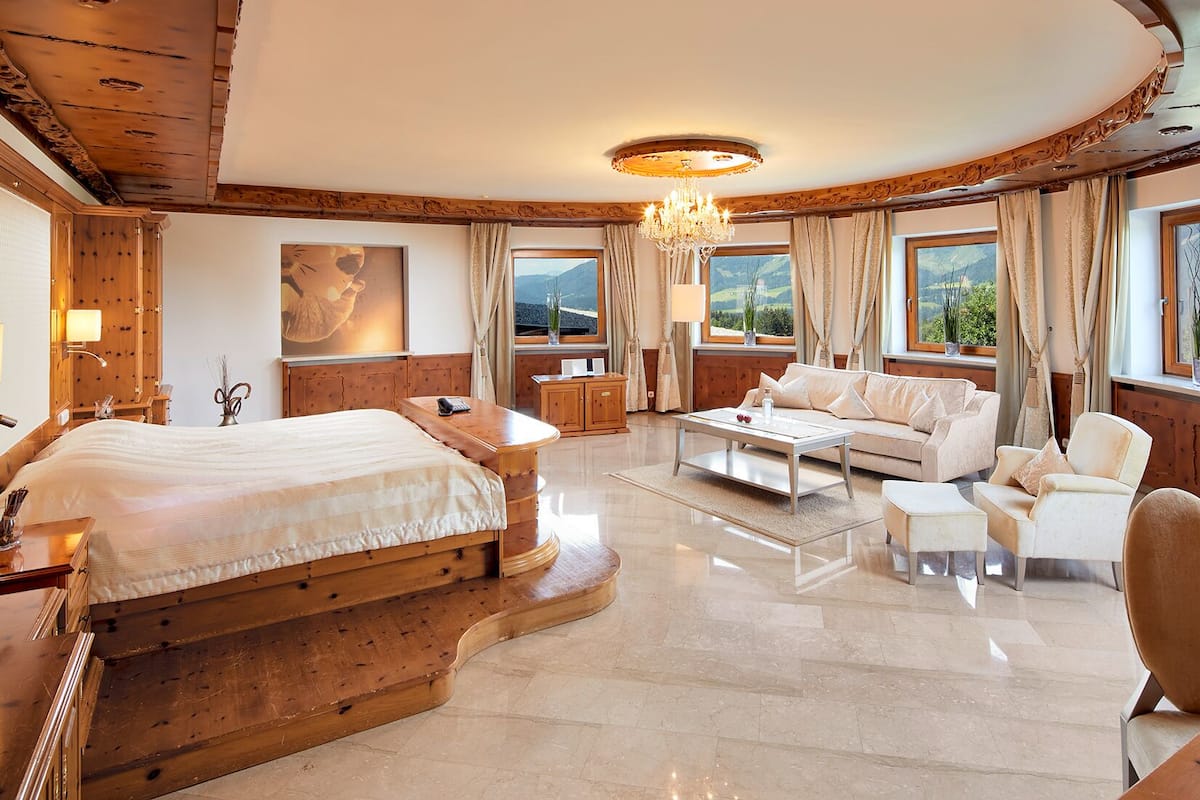 A spacious bedroom with a large bed, a wooden headboard, a desk, a sofa, and a chair. There are multiple windows with curtains, a chandelier, and a painting on the wall.