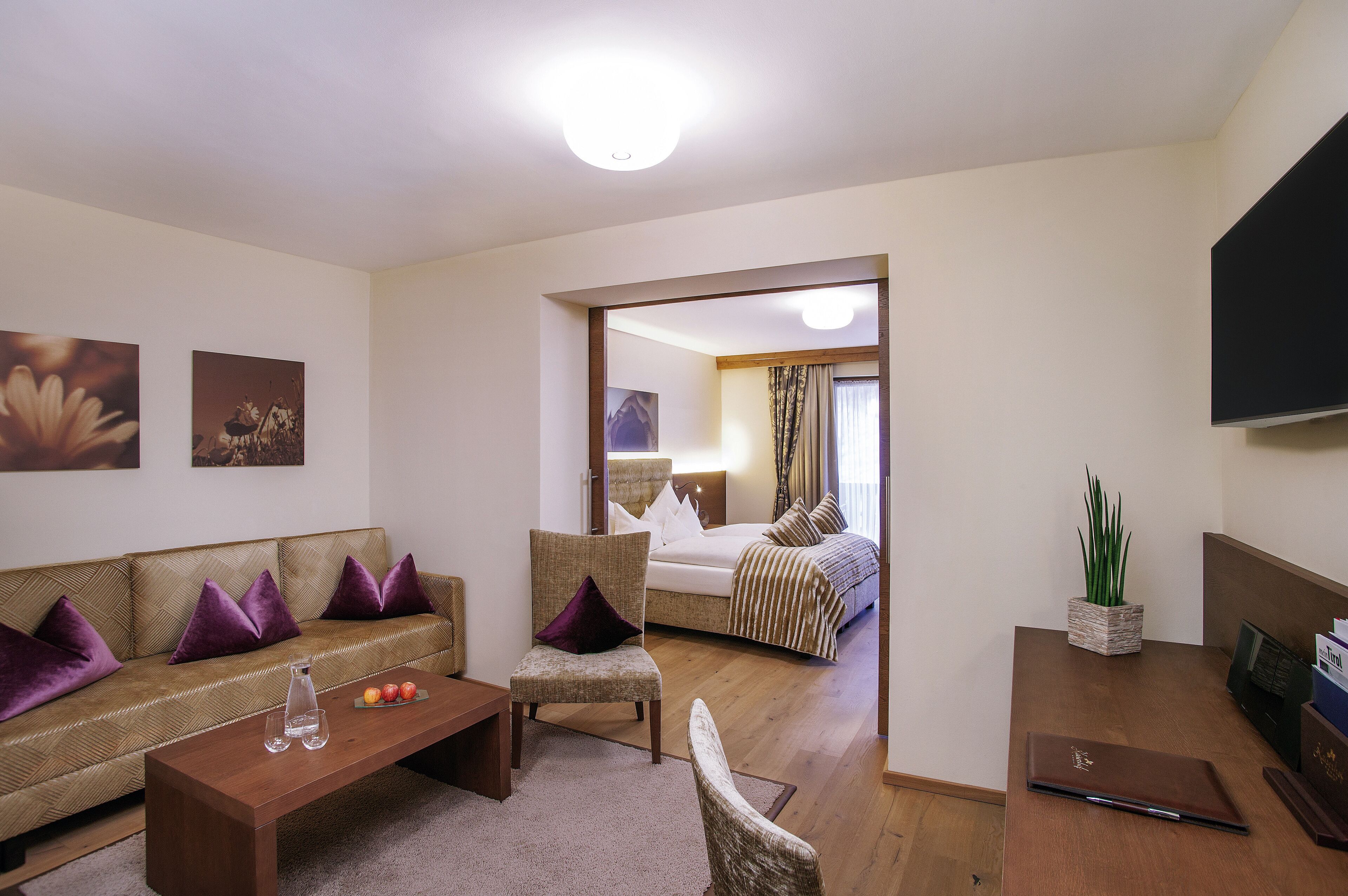 Una habitación de hotel moderna con sofá, sillones, una mesa de centro de madera, una cama con ropa de cama a rayas, un televisor y una planta.