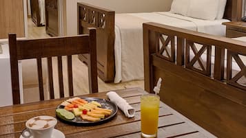 A hotel room with a wooden bed, a dining table with a plate of fruit and a drink, and a chair.