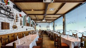 Un restaurant amb taules preparades per dinar, una paret amb noms de ciutats i vista a un cos d'aigua.