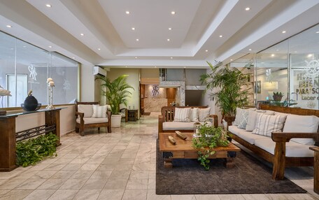 A spacious living area with a wooden sofa, a coffee table, and potted plants. There is a fireplace and a kitchen area in the background.