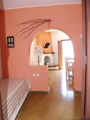 A room with a bed, a small table, a lamp, and a window. There is a door leading to another room. - Cretan Village (Agios Nikolaos)