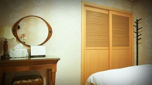 Un dormitorio con un armario de madera, un espejo redondo, una mesita de noche con lámpara y una cama con sábana blanca.