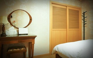 Un dormitorio con un armario de madera, un espejo redondo, una mesita de noche con lámpara y una cama con sábana blanca.