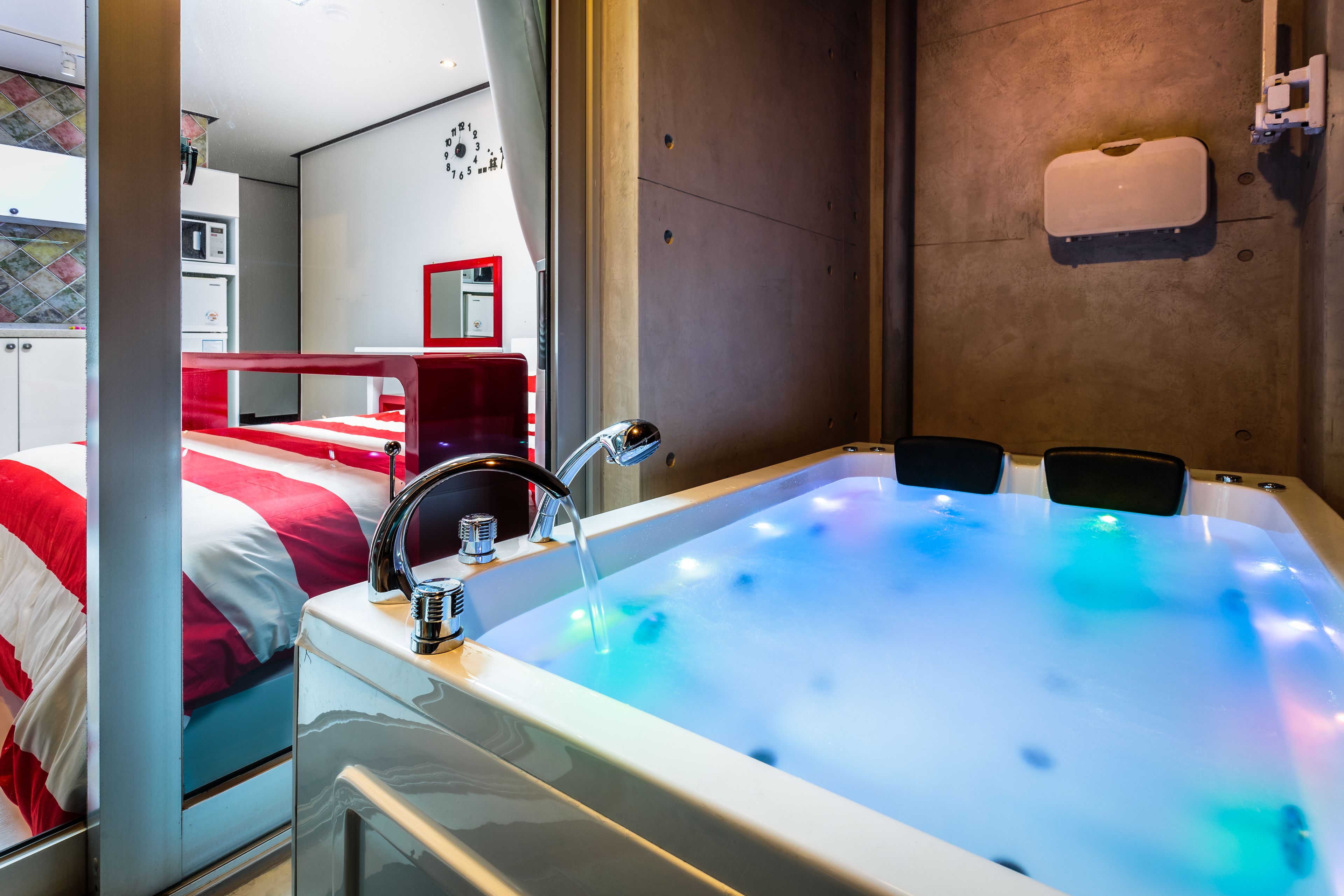 A modern hotel room with a large bathtub, a red bed, and a microwave in the kitchenette.