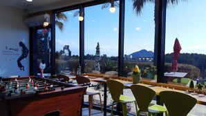 A room with a foosball table, green chairs, and large windows offering a view of a mountain and greenery. - Beewool Chaewool (Seogwipo)