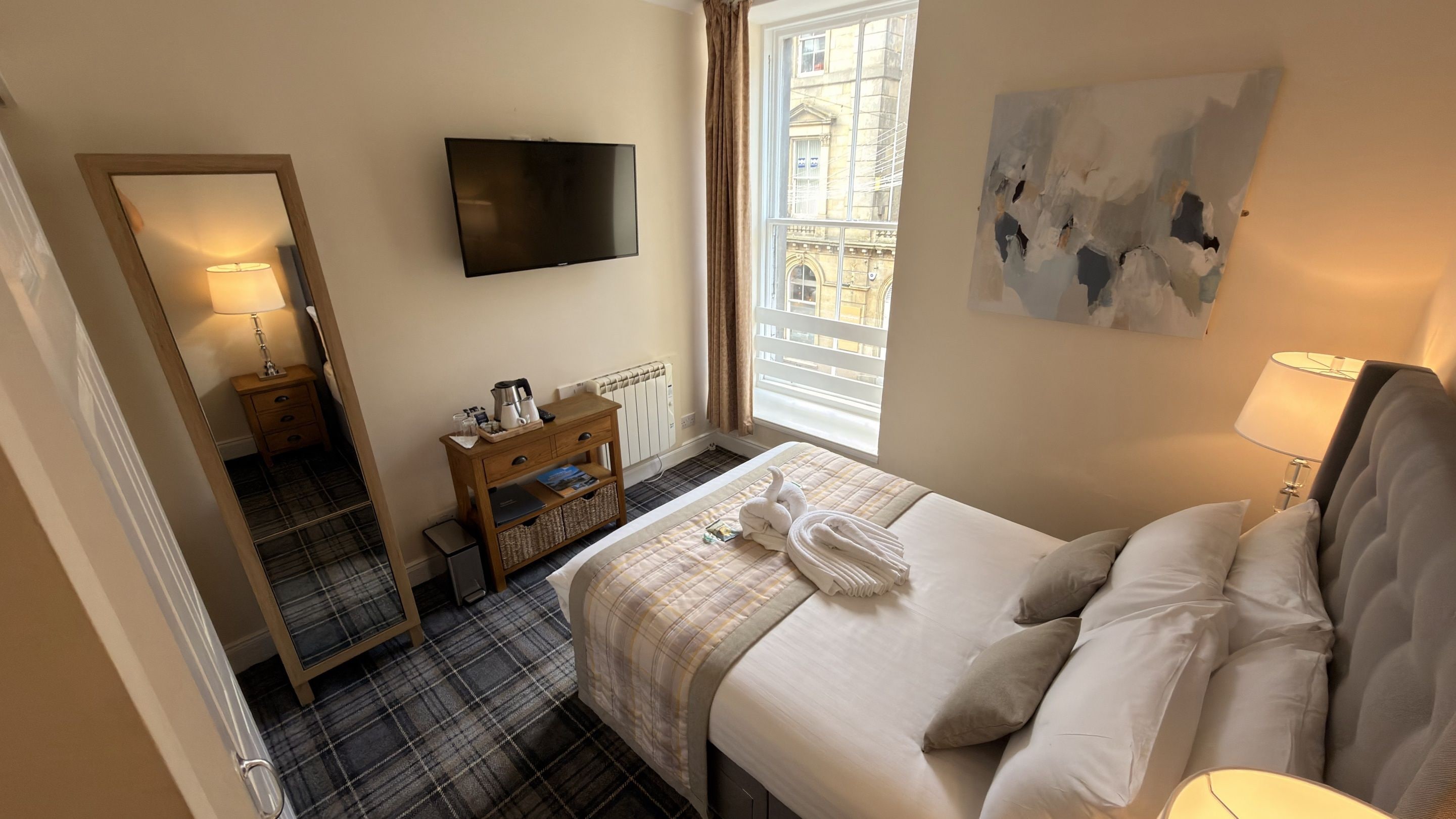 A hotel room with a bed, a flat-screen TV, a window with curtains, a painting on the wall, and a wooden nightstand with a lamp.