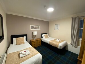 A hotel room with two beds, a nightstand with a lamp, and a window with curtains.