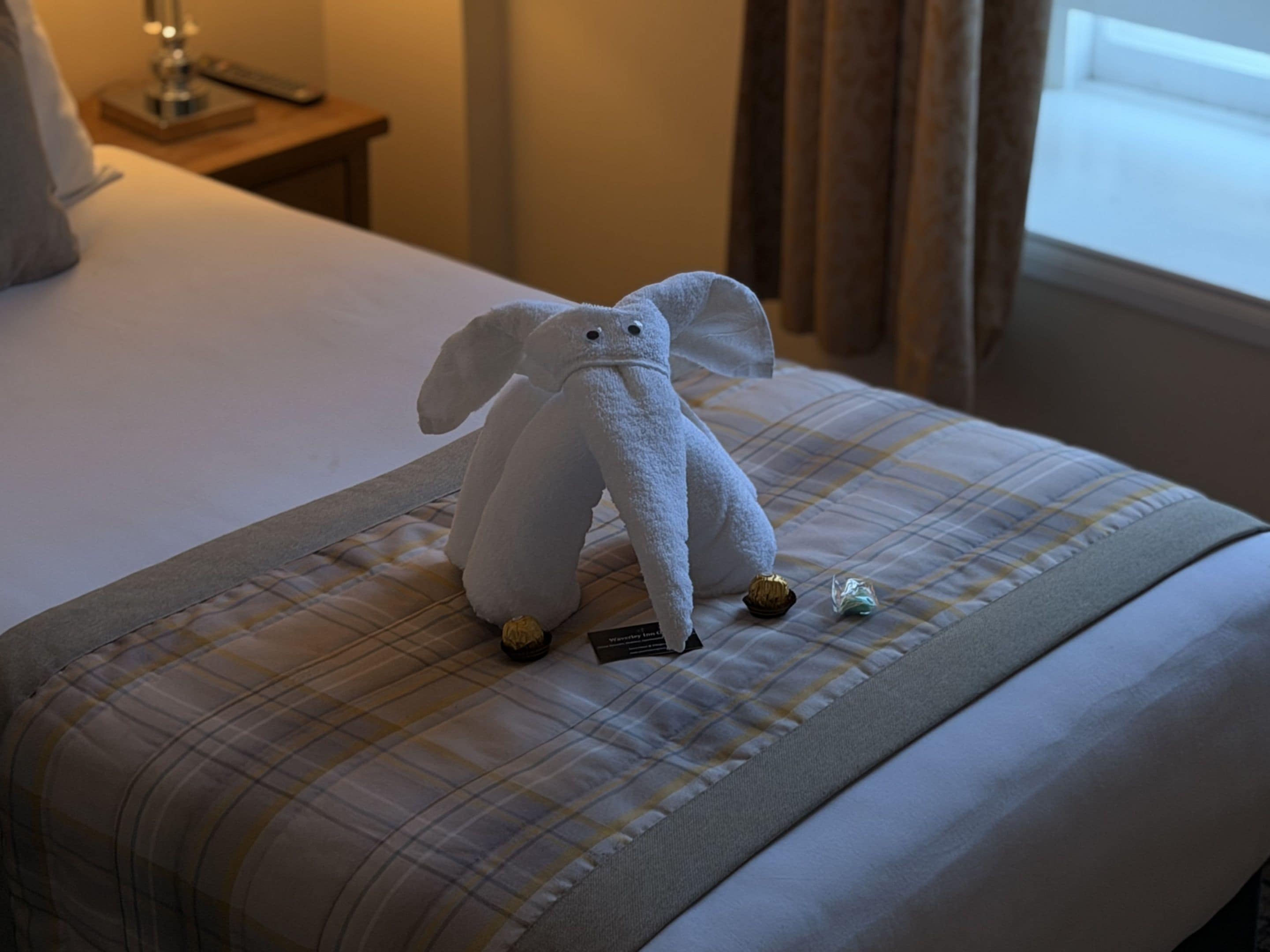 A bed with a plaid bedspread, a white towel elephant, and a small bottle on the bed.