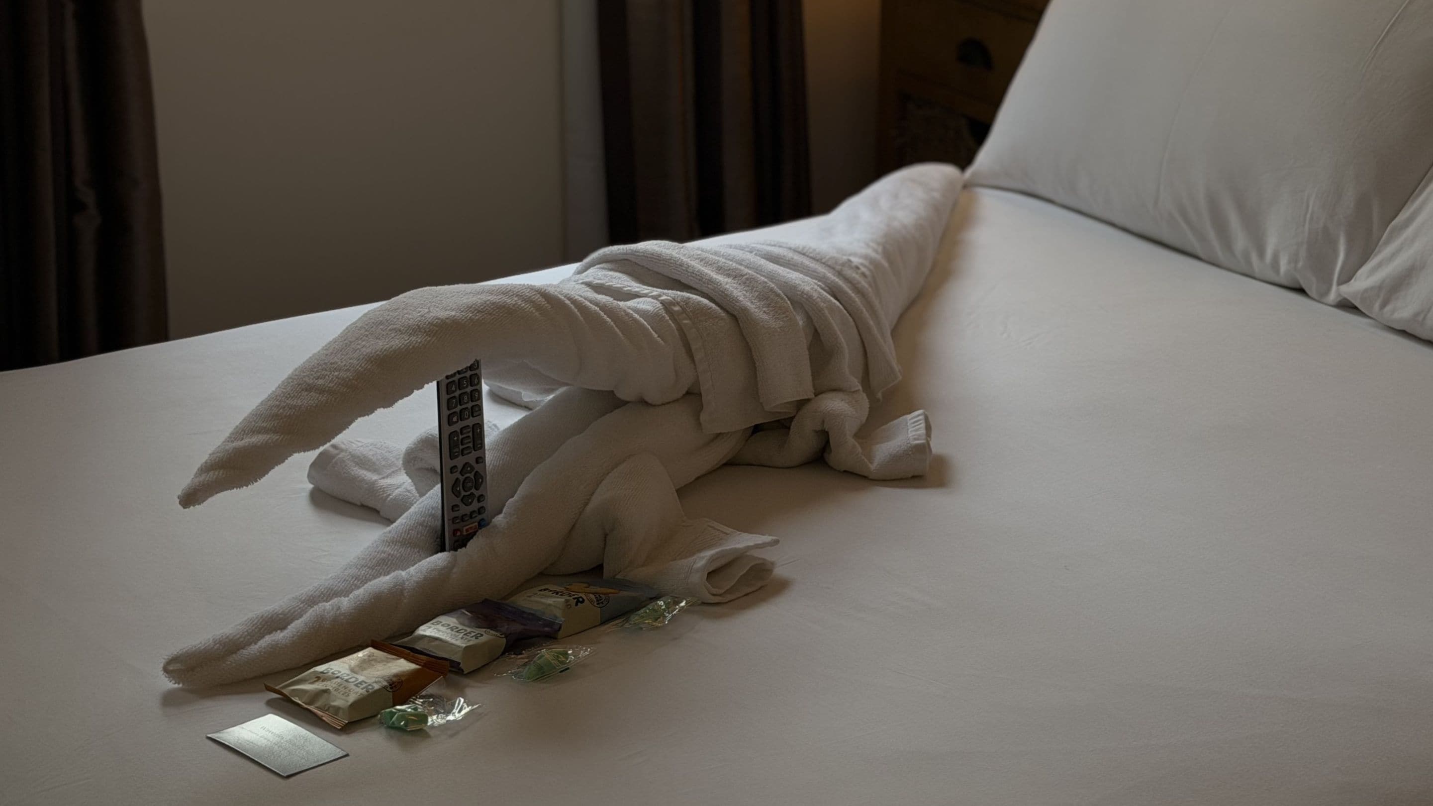A neatly made bed with a folded towel, a remote control, and a few toiletries.