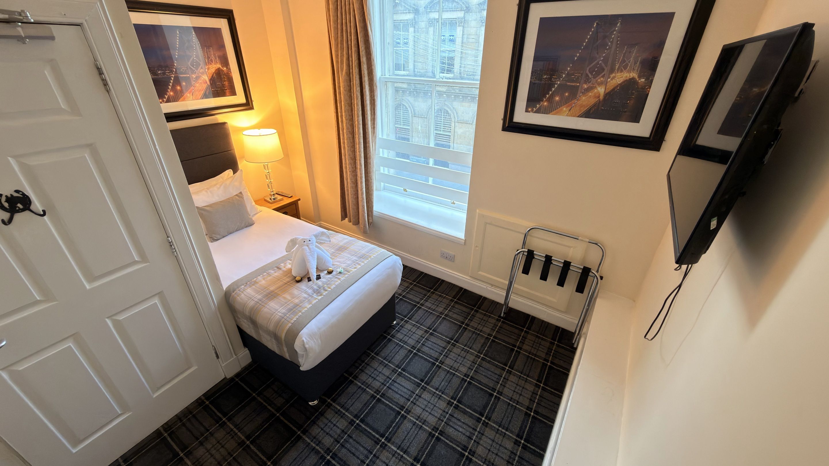 A hotel room with a bed, a bedside lamp, a window with curtains, a wall-mounted TV, and a framed picture of a bridge.
