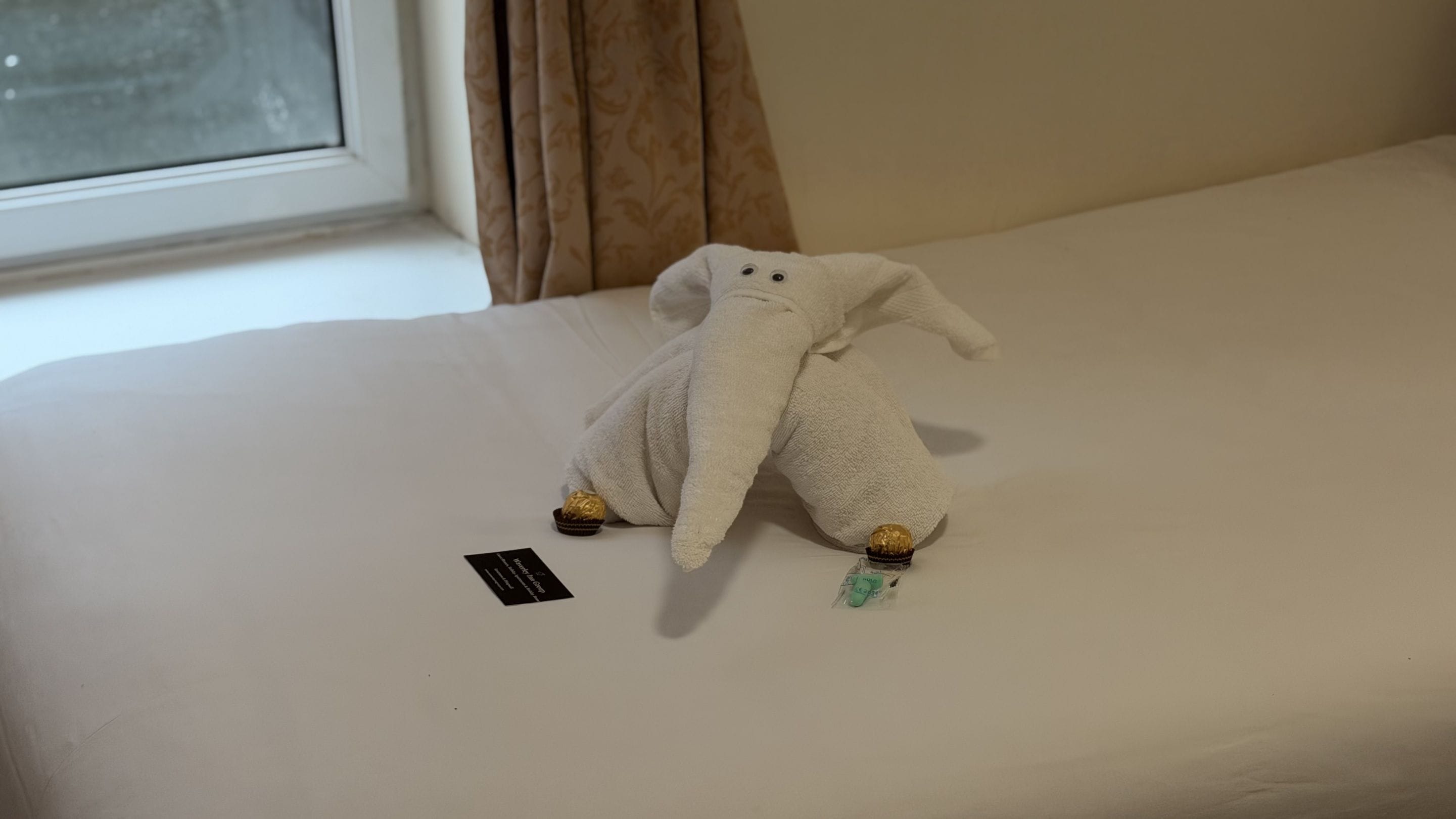 A plush toy elephant on a bed with a black tag and a window with curtains.