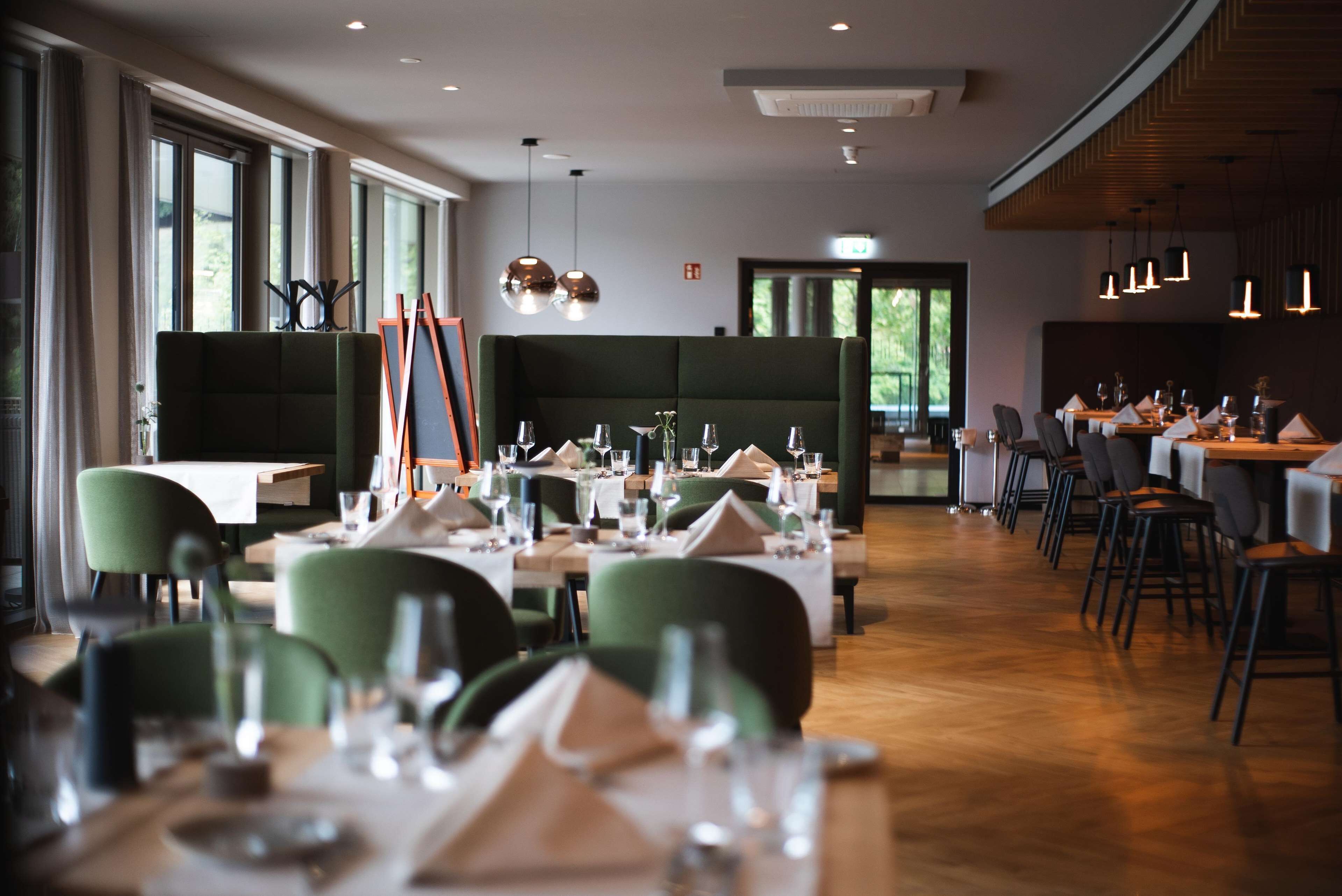 Un restaurante con mesas dispuestas para comer, sillas tapizadas en verde e iluminación colgante.