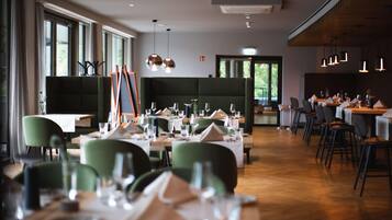 Un restaurante con mesas dispuestas para comer, sillas tapizadas en verde e iluminación colgante.
