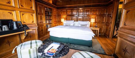 A wooden-paneled room with a large bed, two bedside tables with lamps, a coffee maker, a bookshelf, and two plaid armchairs.
