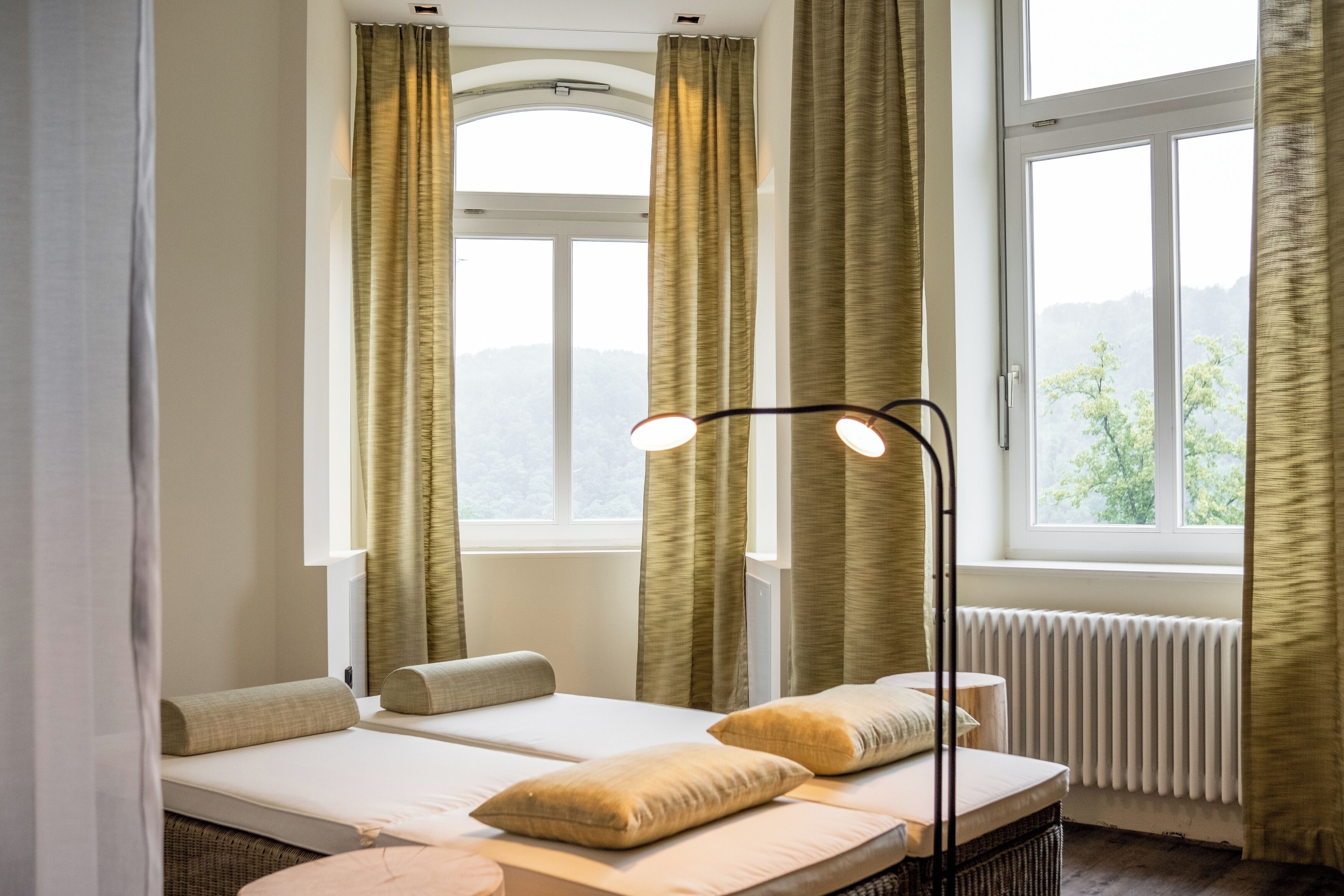 Una habitación con dos camas, un radiador y vistas a árboles por las ventanas.