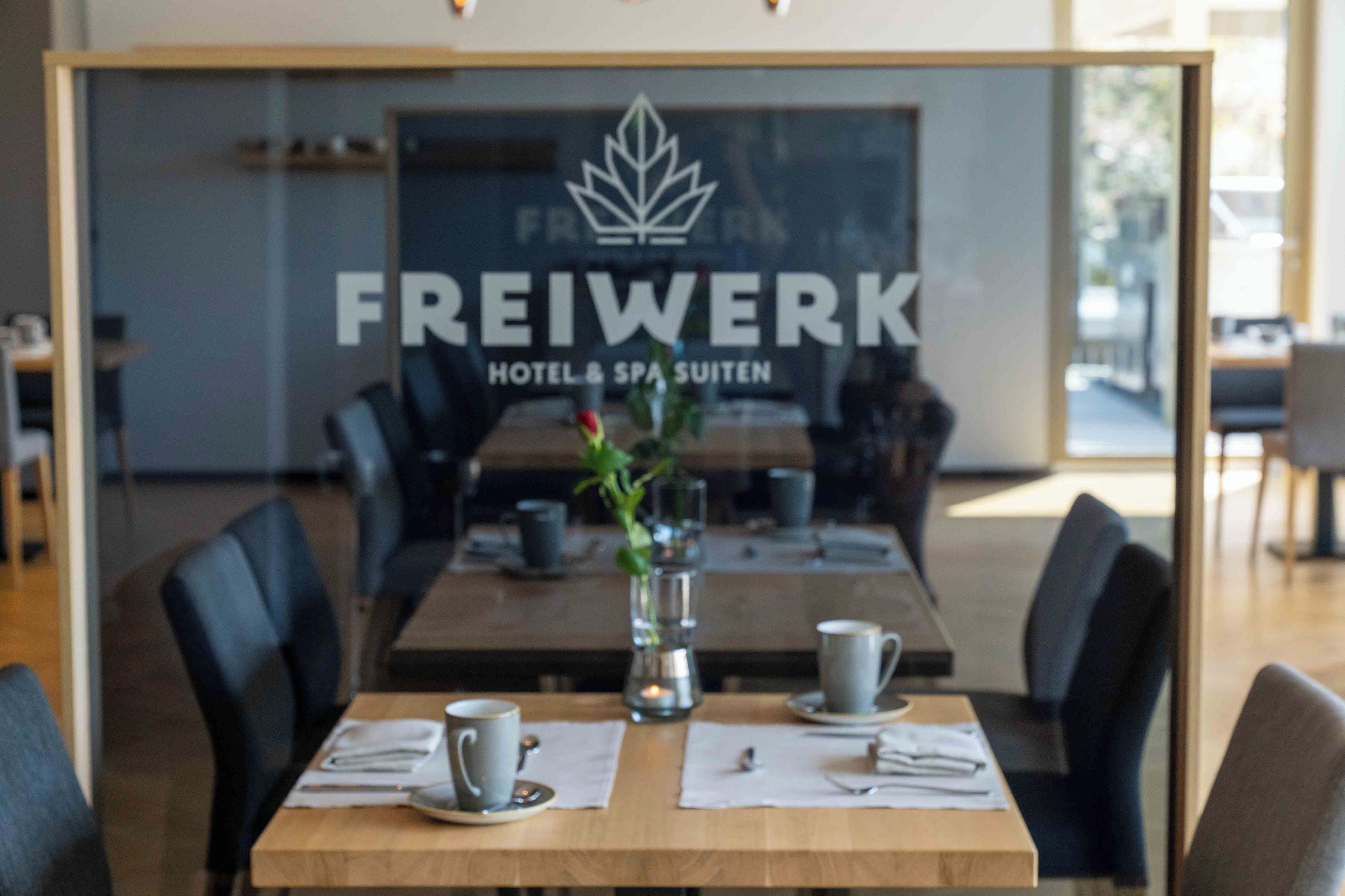 A modern dining area with a wooden table set for two, a glass partition with the text "FREIWERK HOTEL & SPA SUITEN" and a logo, and a vase with a plant.