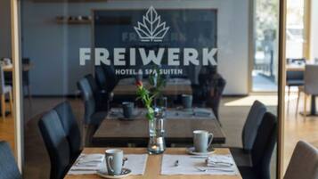 A modern dining area with a wooden table set for two, a glass partition with the text "FREIWERK HOTEL & SPA SUITEN" and a logo, and a vase with a plant.
