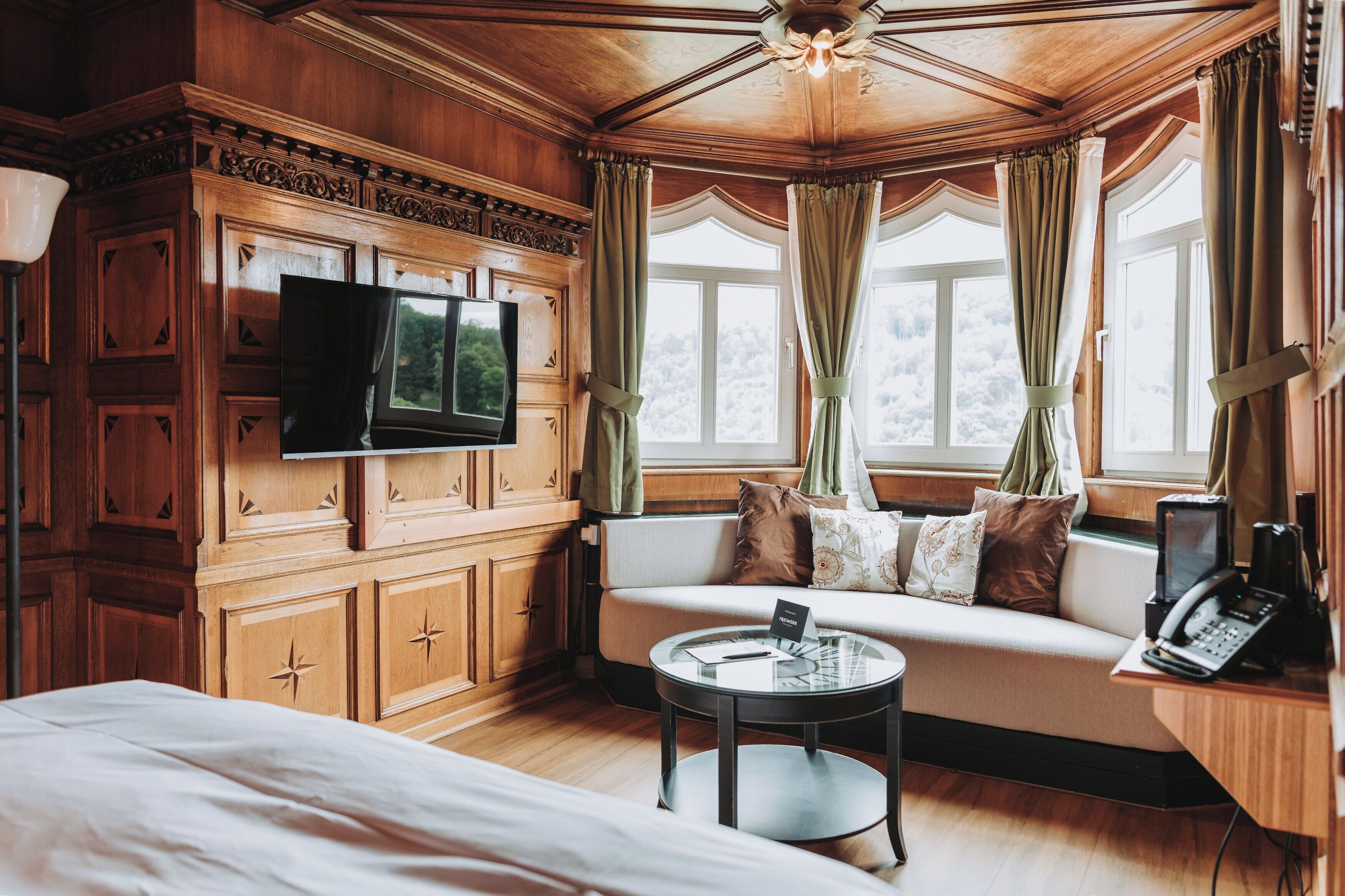 Una habitación con techo de madera, un sofá con cojines, un televisor de pantalla plana montado en un mueble de madera, una mesa redonda de vidrio y un teléfono sobre un escritorio de madera.