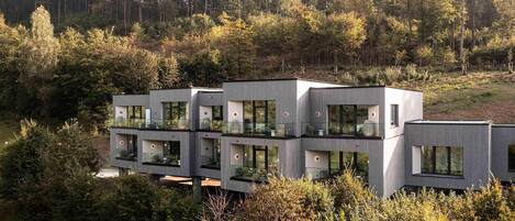 A modern multi-story building with large windows and balconies, surrounded by dense forest.