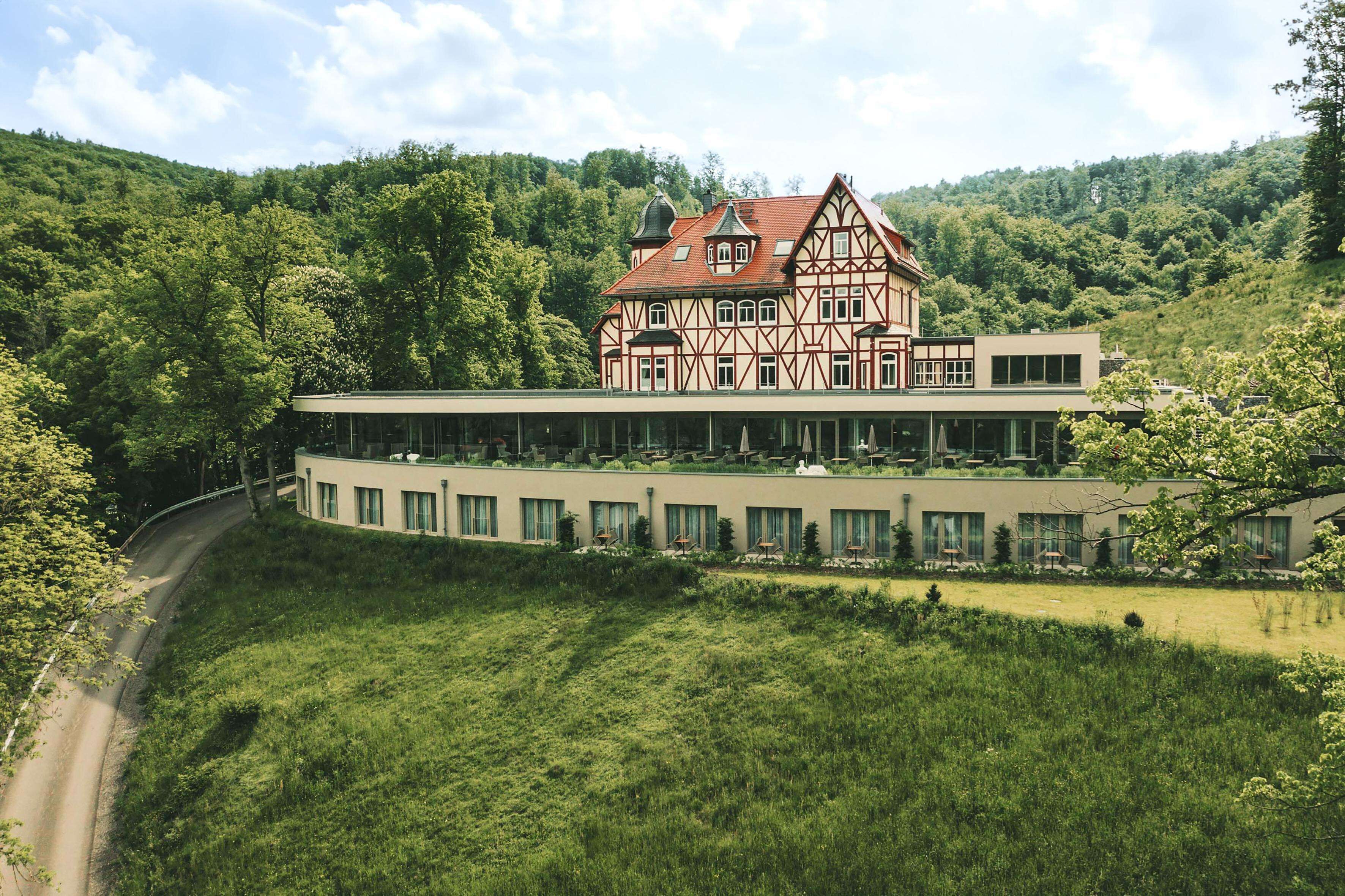 A traditional half-timbered house with a modern annex, surrounded by lush greenery and a winding road.