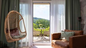 Una habitación con una silla colgante de mimbre, un sofá marrón y vistas a un paisaje verde a través de un ventanal.