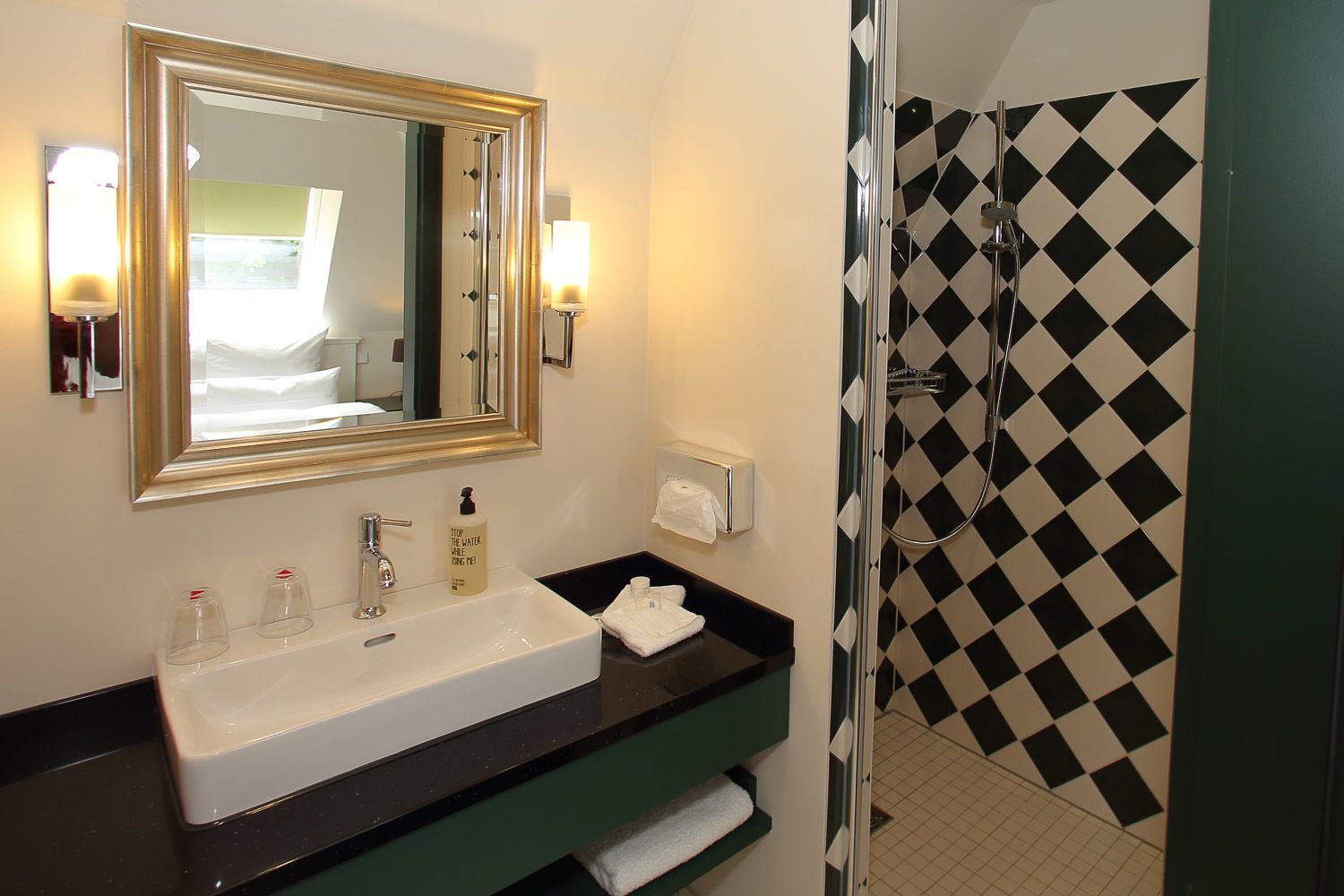 A bathroom with a large mirror, two sinks, a shower with a black and white checkered wall, and a green vanity.