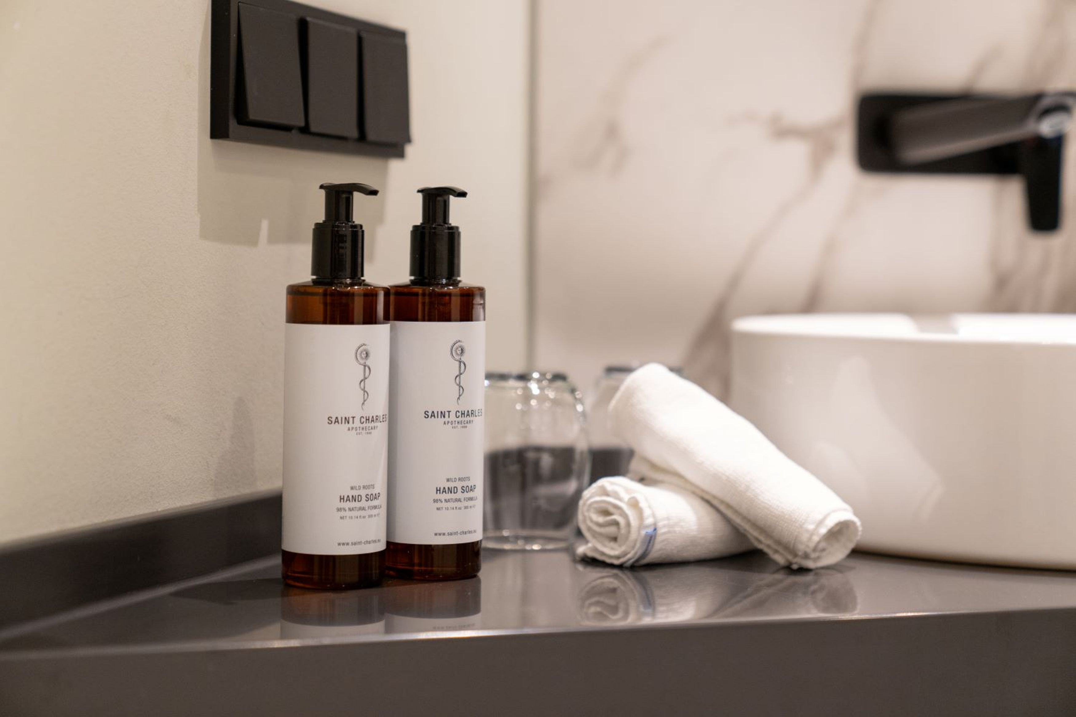 A bathroom with two bottles of Saint Charles hand soap, a glass, and a rolled white towel on a countertop.
