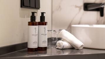 A bathroom with two bottles of Saint Charles hand soap, a glass, and a rolled white towel on a countertop.