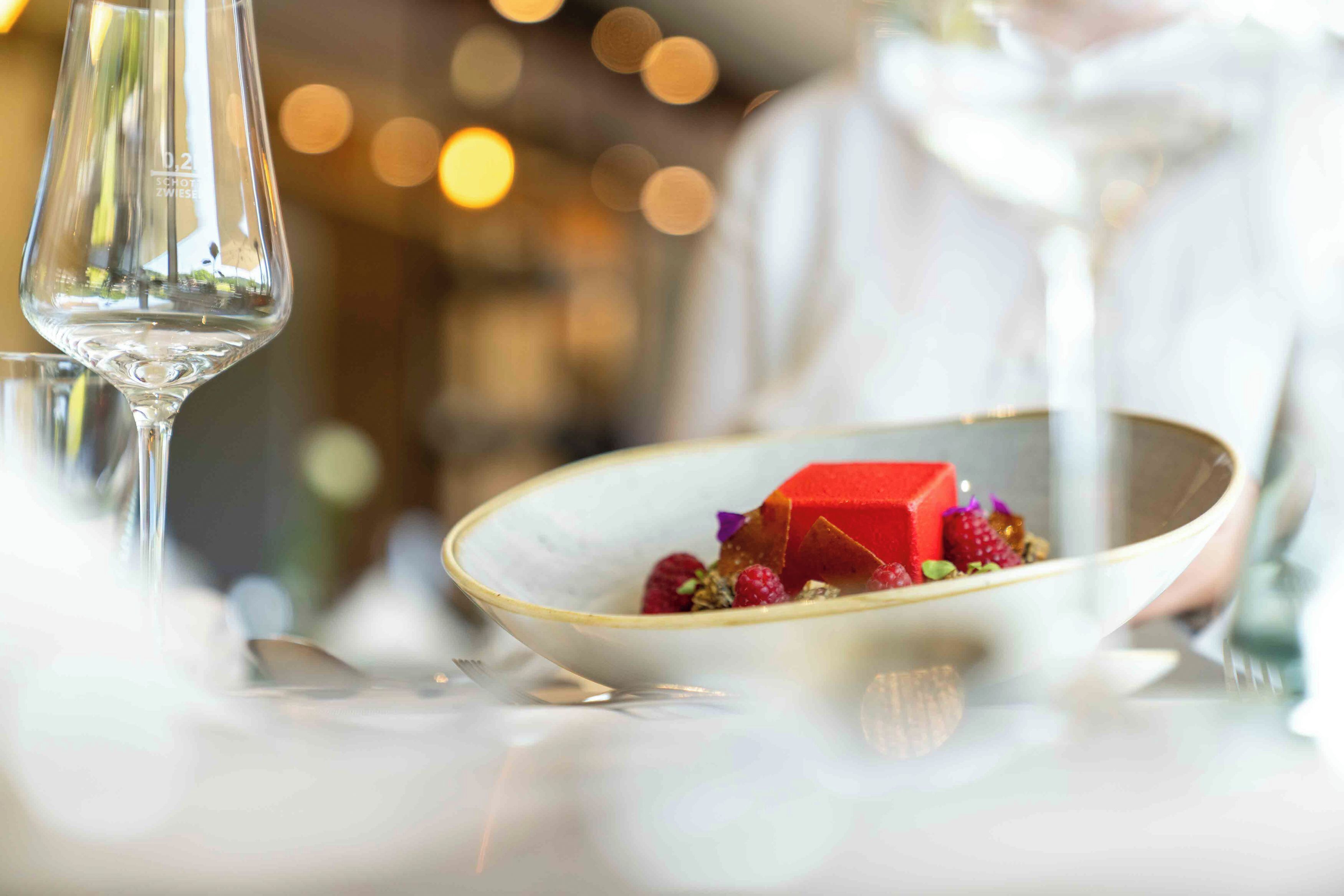 Un plato de postre con un cubo rojo y bayas, una copa de vino y un fondo desenfocado con iluminación cálida.