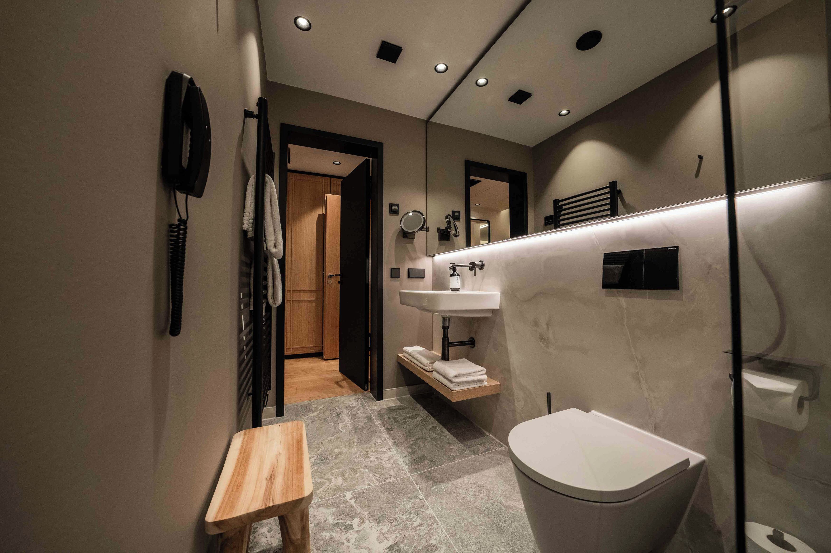 A modern bathroom with a toilet, sink, and mirror. There is a wooden bench and a towel rack.