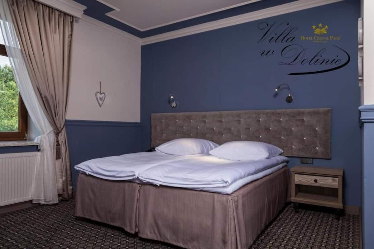 Une chambre à coucher comprenant un lit, une table de chevet, une lampe fixée au mur et une fenêtre avec des rideaux.