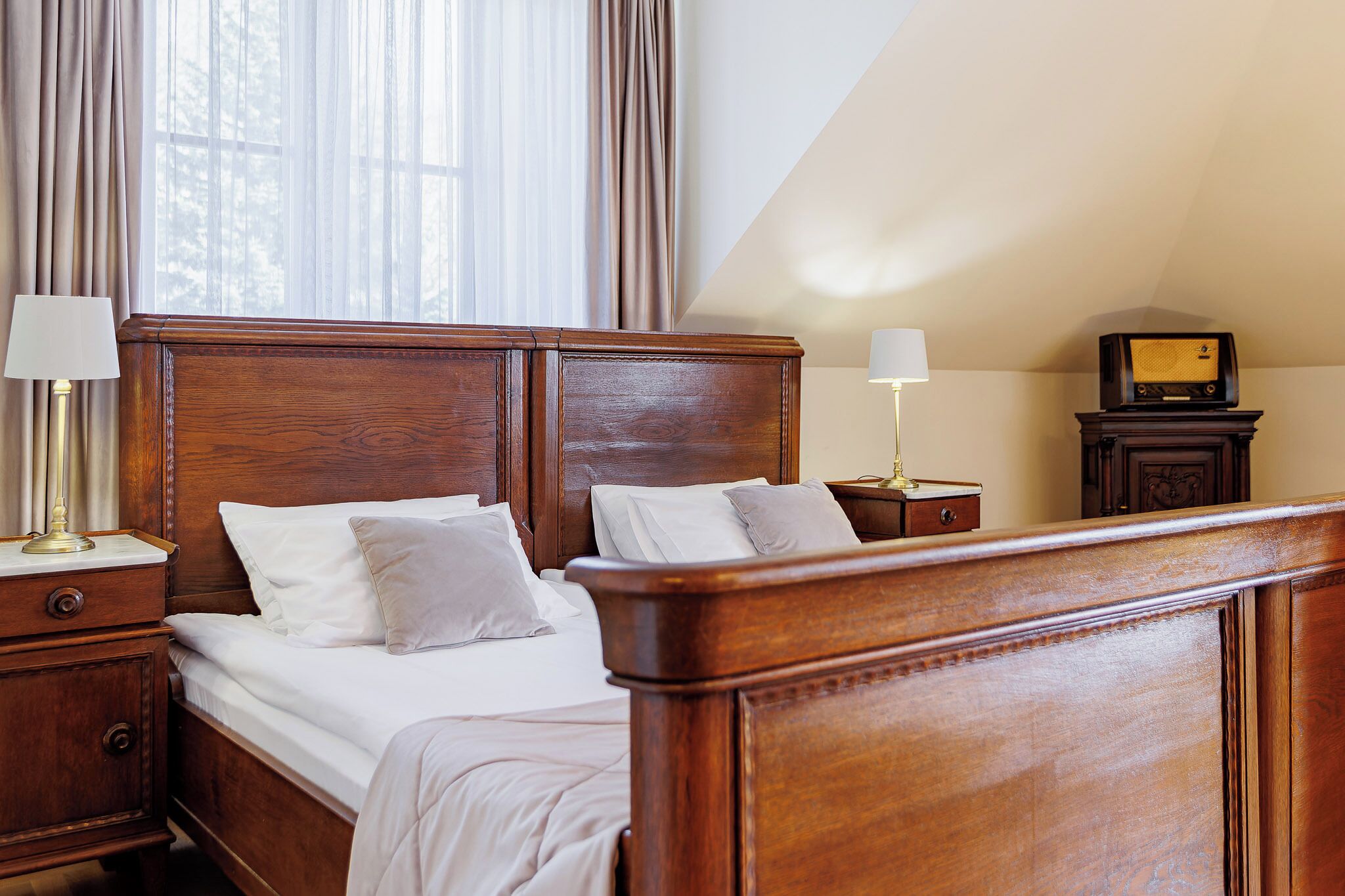 A neatly made bed with white linens and two pillows, a wooden headboard, a bedside table with a lamp, and a radio on a small table beside the bed.