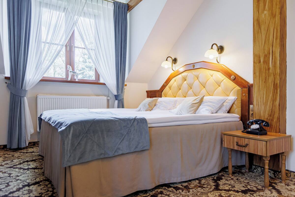 Un lit bien fait, avec une tête de lit, une table de chevet avec une lampe et une fenêtre ornée de rideaux légers.