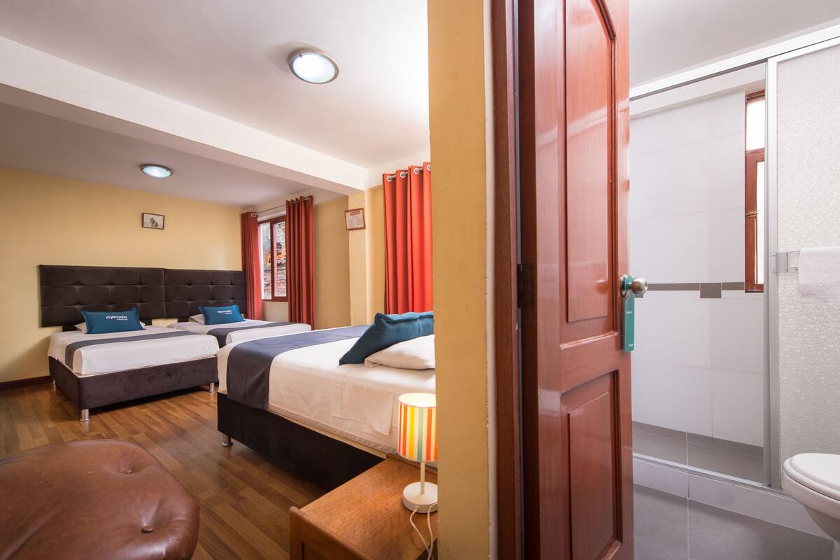 A hotel room with two beds, a wooden bench, a lamp, and a bathroom visible through an open door.