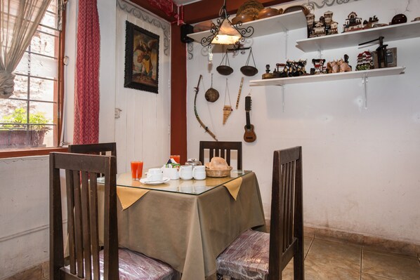 A dining area with a table set for two, chairs, a hanging light fixture, and shelves with decorative items.