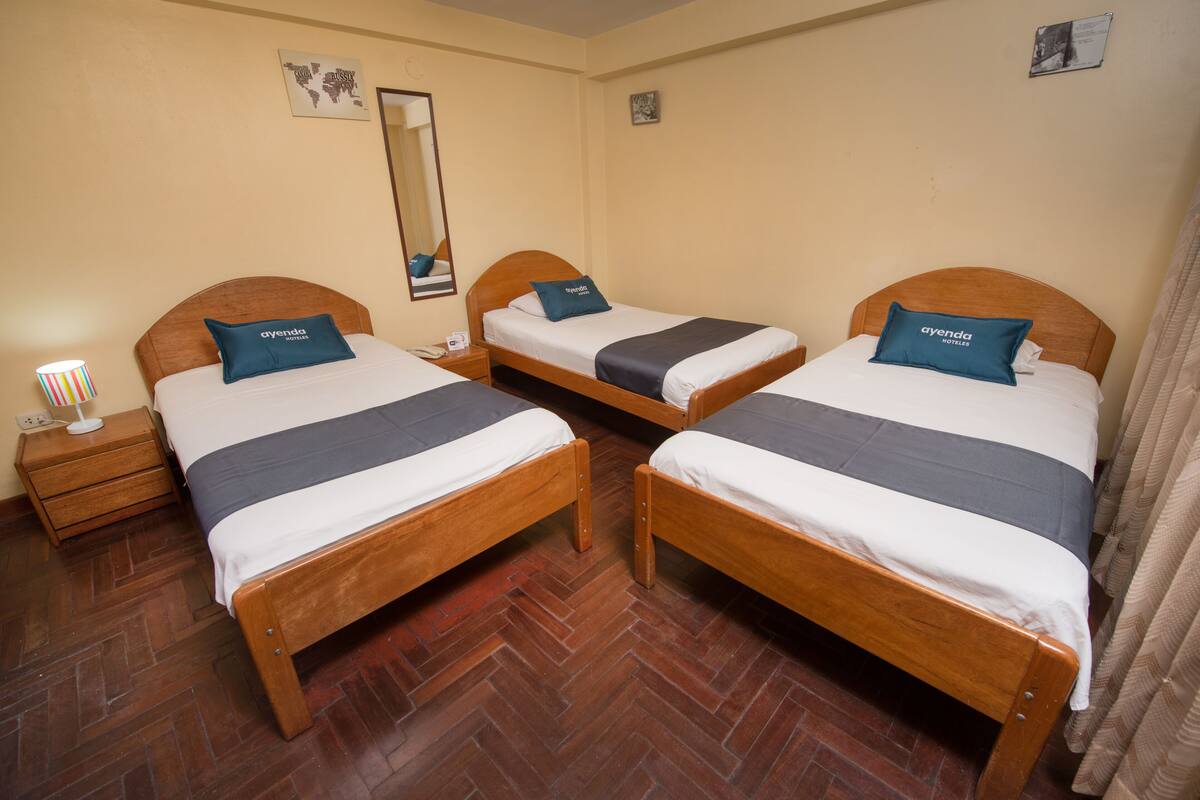 A hotel room with three wooden beds, each with a blue pillow and white bedding. There is a wooden nightstand with a lamp and a phone on it. The room has a herringbone-patterned wooden floor and beige walls.